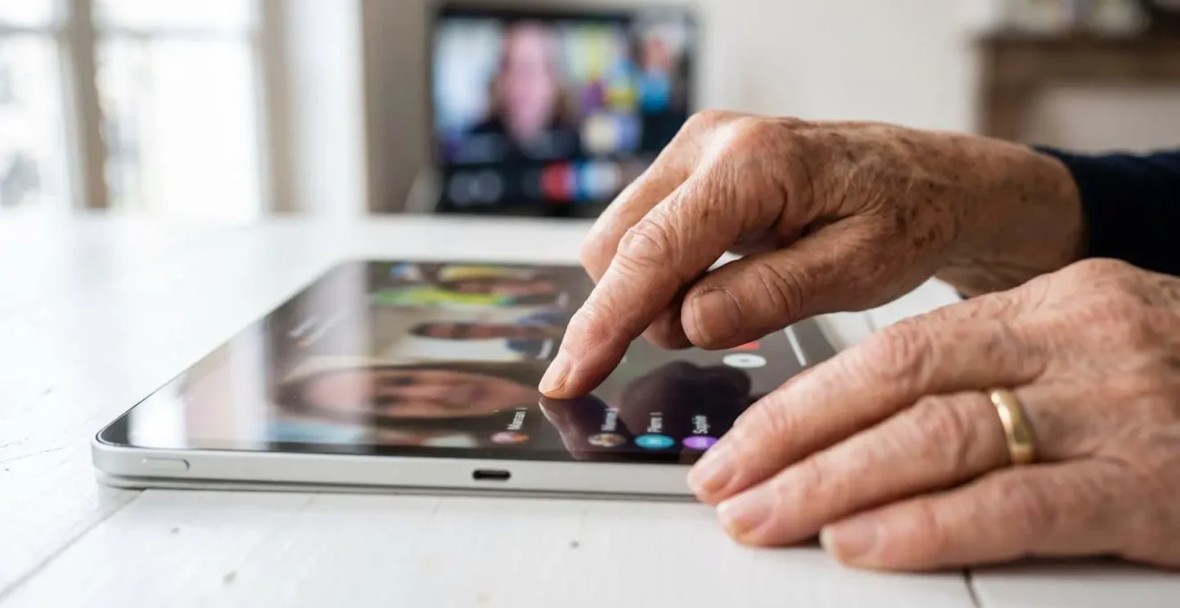 Gros plan sur des mains de personne âgée utilisant une tablette tactile avec un geste doux sur l'écran, arrière-plan flouté montrant une interface de visioconférence